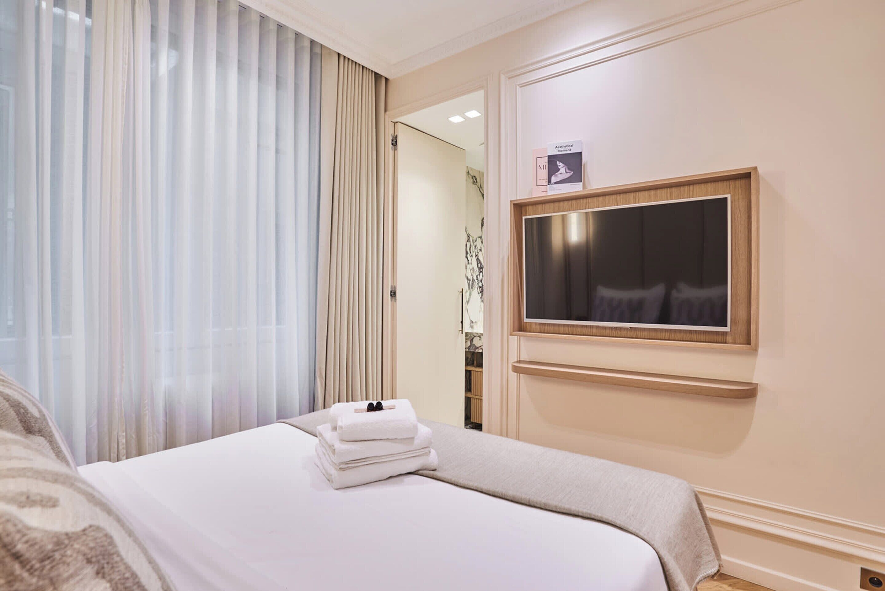 A hotel room with a bed, a TV mounted on the wall, and a window with sheer curtains.
