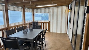 Una terraza cubierta al aire libre con una mesa, sillas, barandales de madera y ventanas amplias.