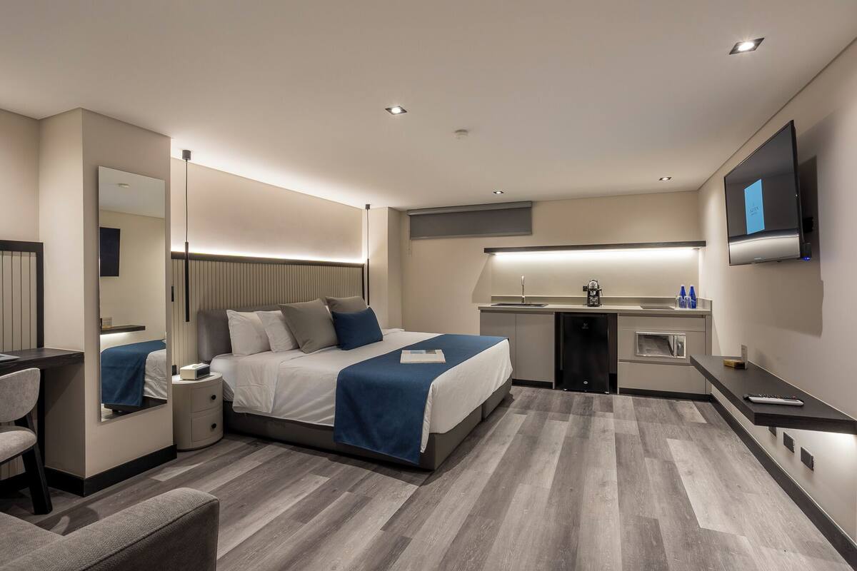 A modern hotel room with a large bed, bedside tables, a desk, a mirror, a TV, and a kitchenette.