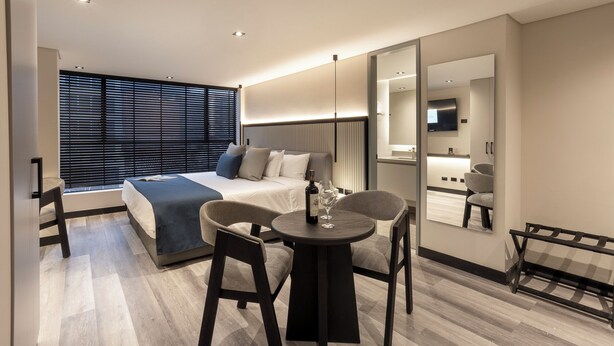 Et moderne hotelværelse med en stor seng, et siddeområde med to stole, et lille bord og et stort spejl.