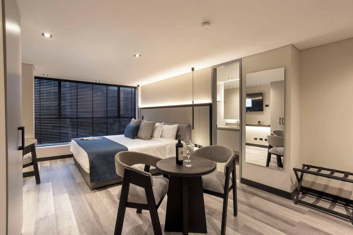 A modern hotel room with a large bed, a seating area with two chairs, a small table, and a large mirror.