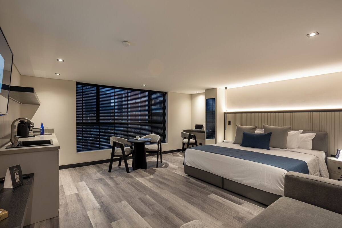 A modern hotel room with a large bed, a desk, and a chair. There is a window with blinds, a mirror, and a small table with a lamp.