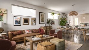 En moderne stue med sofa, sofabord og skærmstole, et spiseområde med stole, samt et køkken med skabe og vask.