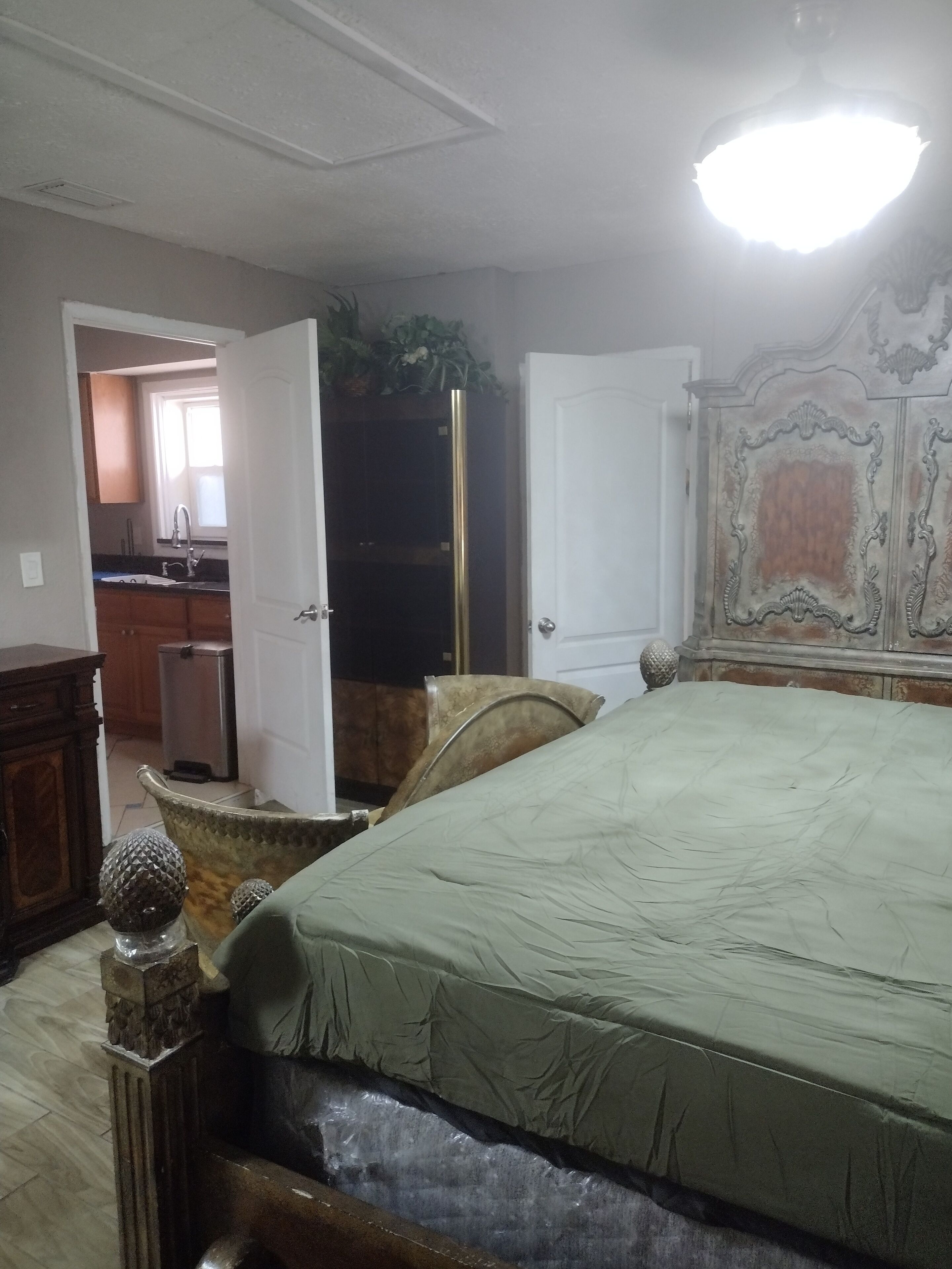 A bedroom with a bed, a wardrobe, a mirror, and a small kitchenette in the background.