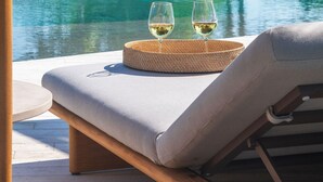 Μια πολυθρόνα χαλάρωσης με ένα πλεκτό τραπεζάκι και δύο ποτήρια λευκό κρασί, με θέα μια πισίνα και πλούσια βλάστηση.