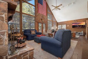 A spacious living room with large windows, a stone fireplace, and a ceiling fan. - Pah's Haus | Hot Tub | Scenic View | Dog-Friendly (Angel Fire)