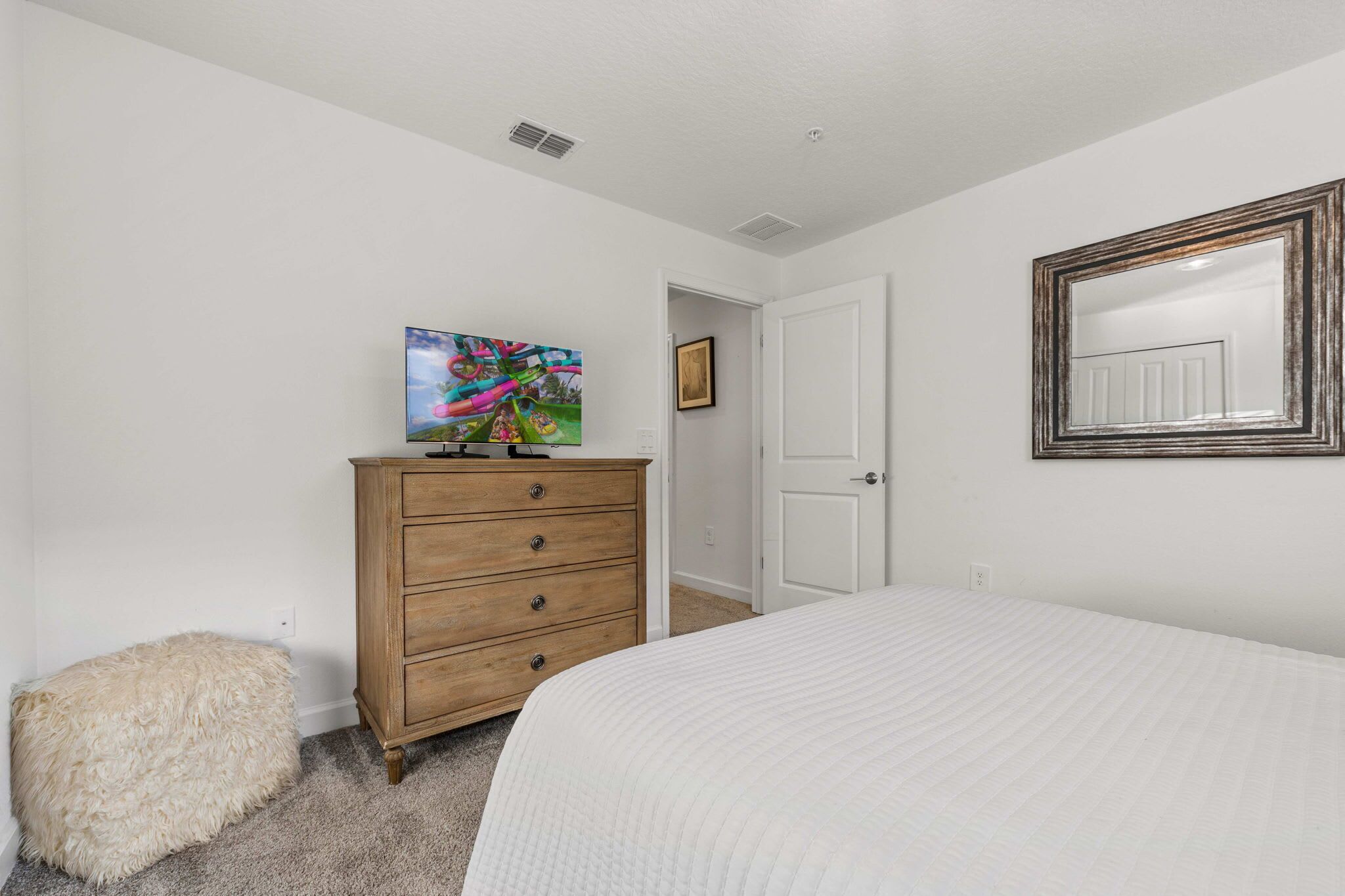 Une chambre à coucher avec un lit, une commode en bois, un pouf moelleux, un tableau et un miroir au mur.