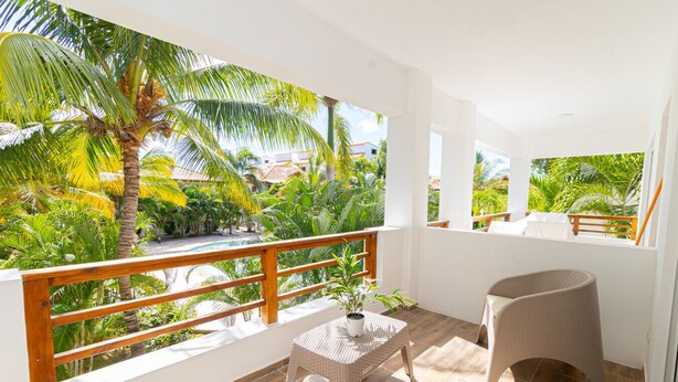 A balcony with a wooden railing, wicker furniture, and a view of a tropical garden with palm trees. - Luxury 1BR Paraiso Bayahibe · Pool · Beach · AC · Kitchen (Bayahíbe)