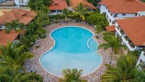 An aerial view of a resort with a large, curved swimming pool surrounded by palm trees and buildings with red-tiled roofs. - Luxury 1BR Paraiso Bayahibe · Pool · Beach · AC · Kitchen (Bayahíbe)