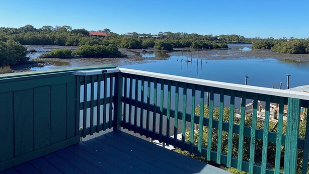 A wooden deck with a railing overlooking a calm water body, surrounded by dense greenery and distant buildings. - Welcome to The Crow's Nest at Fisherman's Wharf, a cozy waterfront 1 bedroom. (Cedar Key)