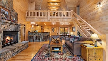 A spacious log cabin interior with a stone fireplace, a dining area, a living room with a sofa and coffee table, and a staircase leading to an upper level.