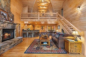 Un intérieur spacieux de chalet en bois, avec une cheminée en pierre, un coin repas, un salon doté d’un canapé et d’une table basse, et un escalier menant à un niveau supérieur.