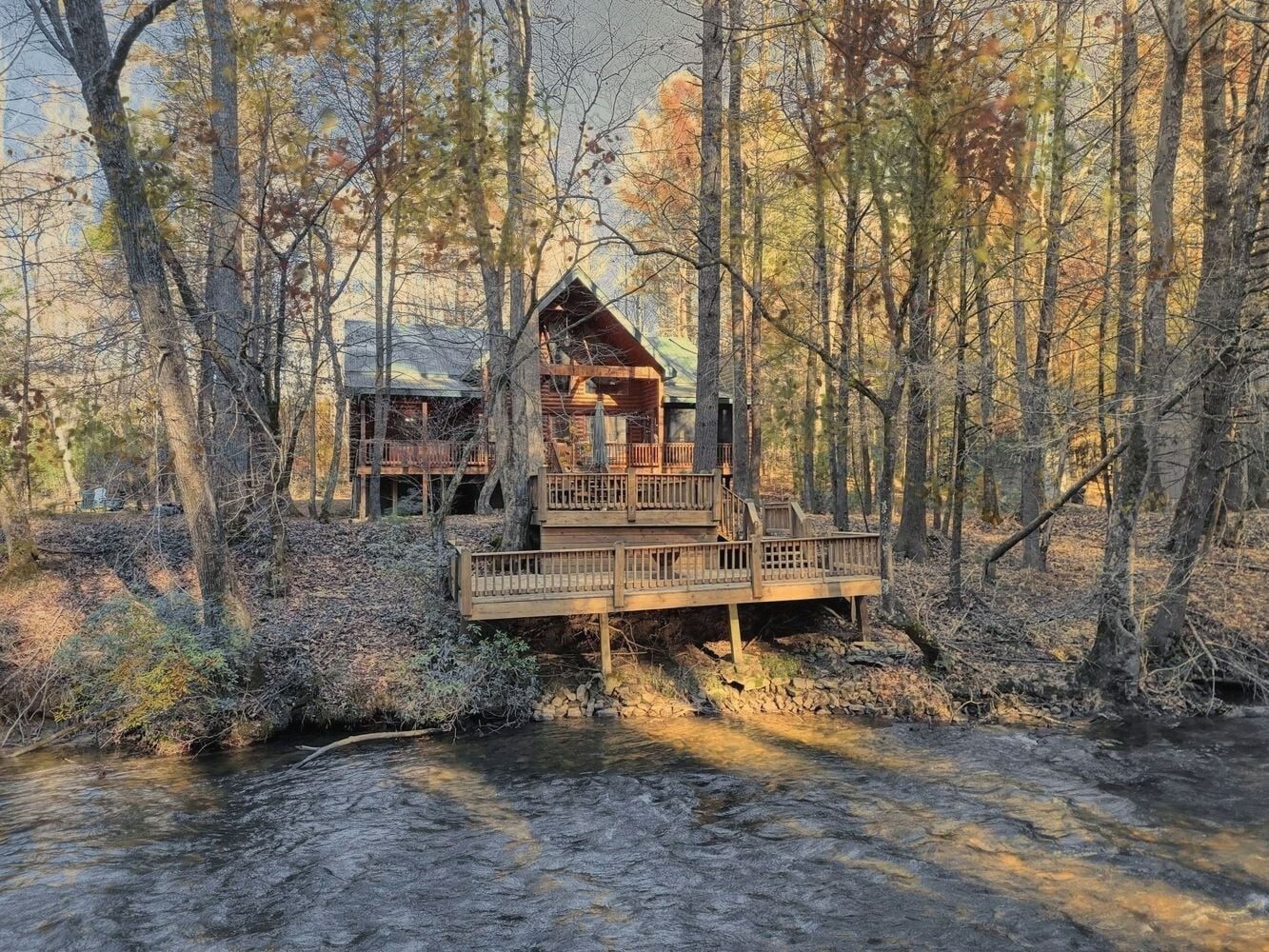 A cabin with a wooden deck and a small bridge over a stream, surrounded by trees.