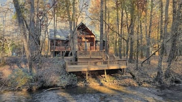 A cabin with a wooden deck and a small bridge over a stream, surrounded by trees.