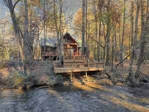 Een chalet met een houten terras en een kleine brug over een beek, omringd door bomen.