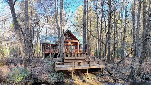 A cabin with a wooden deck and a small bridge over a stream, surrounded by trees. - Tranquility on the Toccoa River - Dog-friendly, 4BR cabin w/hot tub in Suches! (Suches)