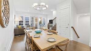 A spacious dining area with a wooden table set for six, surrounded by wooden chairs. A chandelier hangs from the ceiling, and a staircase is visible in the background. - Seacrest, Steps to Beach & Pools! 4 Bikes Included, Pet Friendly! (Rosemary Beach)