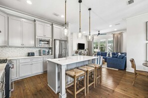 A modern kitchen with white cabinetry, a central island with bar stools, and a dining area with a blue sofa. - Outdoor Oasis! Walk 2 Shops & Eat! LSV/Golf Cart, 3 Kings & Bikes! (Santa Rosa Beach)