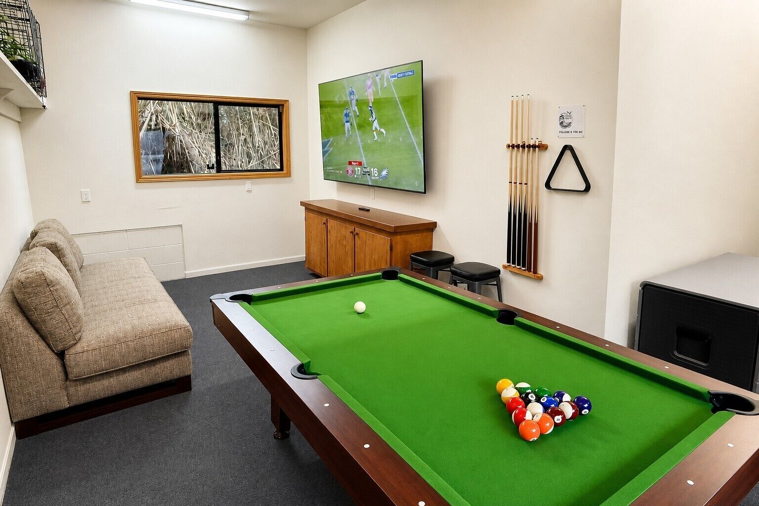 Mesa de bilhar com bolas, sofá, televisão exibindo uma partida esportiva e janela com vista para árvores.