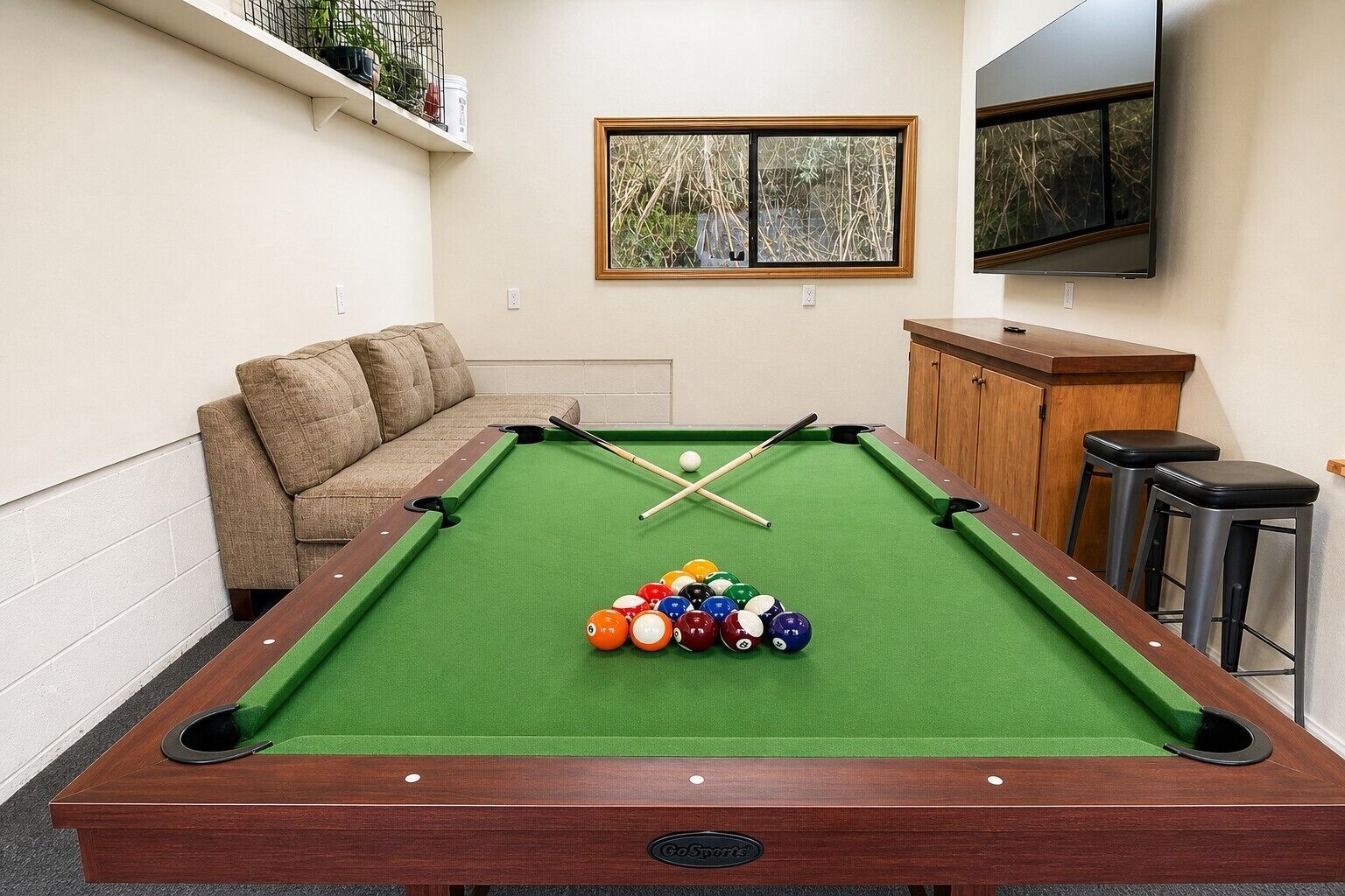 A pool table with balls, a sofa, a TV, and a window with a view of trees.