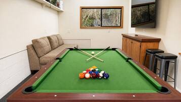 A pool table with balls, a sofa, a TV, and a window with a view of trees.