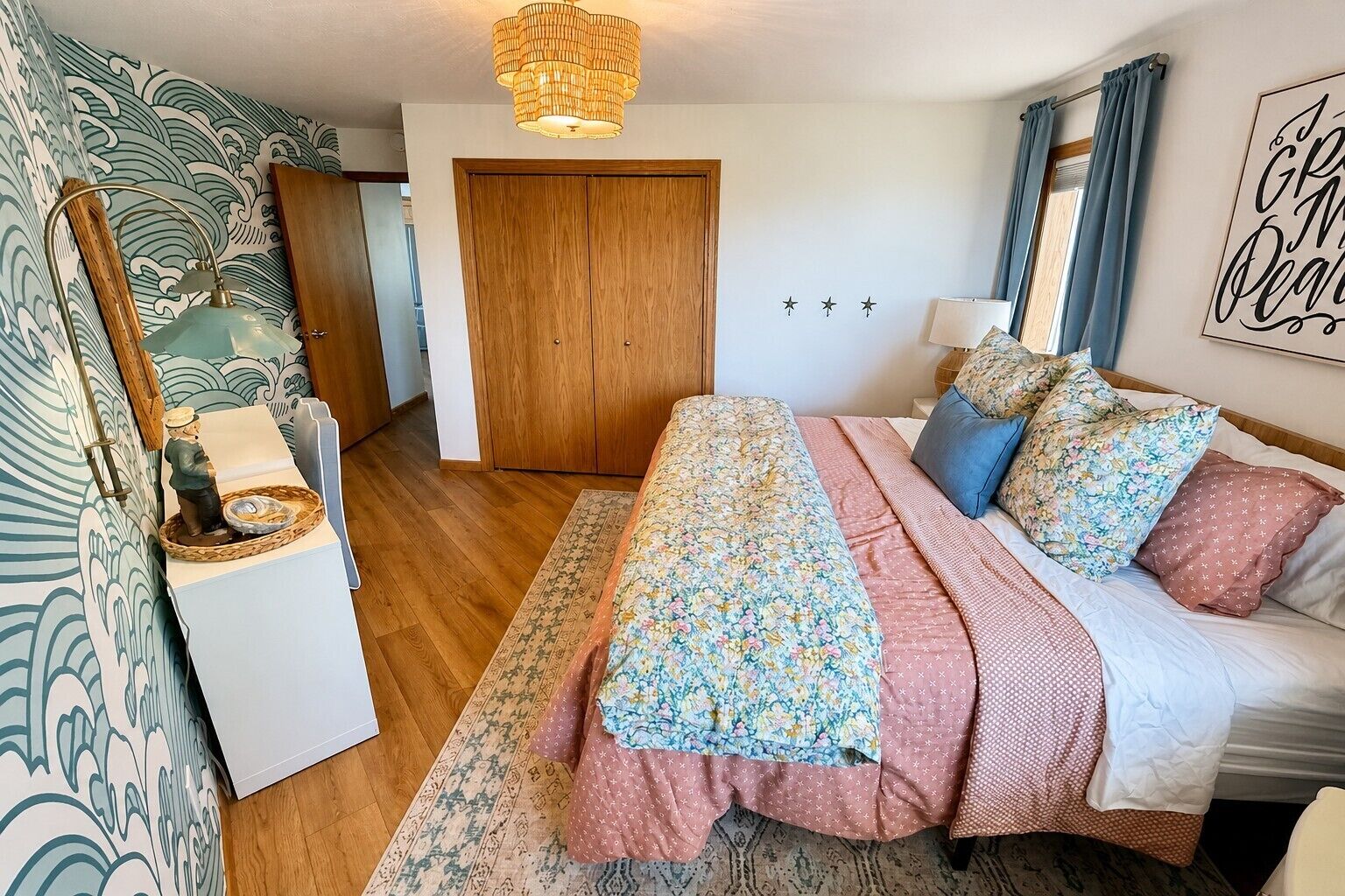 A bedroom with a bed, a wooden wardrobe, a nightstand, a lamp, and a wall with a decorative sign.