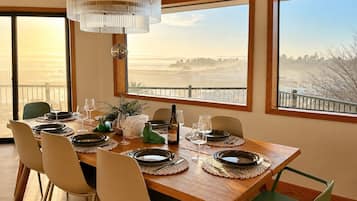 A dining area with a wooden table set for four, large windows with a view, and a chandelier.