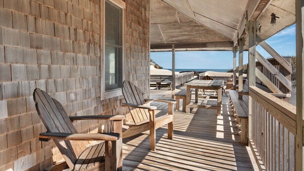 Terraza de madera con sillas Adirondack y mesas de picnic, con vista a una playa.