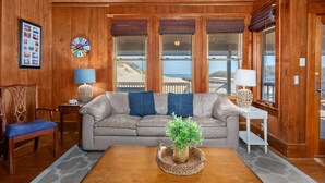 Una sala de estar acogedora con techo de madera, un sofá beige, cojines azules, una mesita de centro con una planta y grandes ventanales con vistas a una playa.