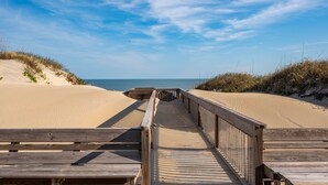 Un paseo marítimo de madera que conduce a una playa con dunas de arena y vegetación escasa.