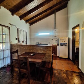 A kitchen with a wooden table, chairs, a refrigerator, and a window with curtains. - Hostal La Merced (Villa General Belgrano)
