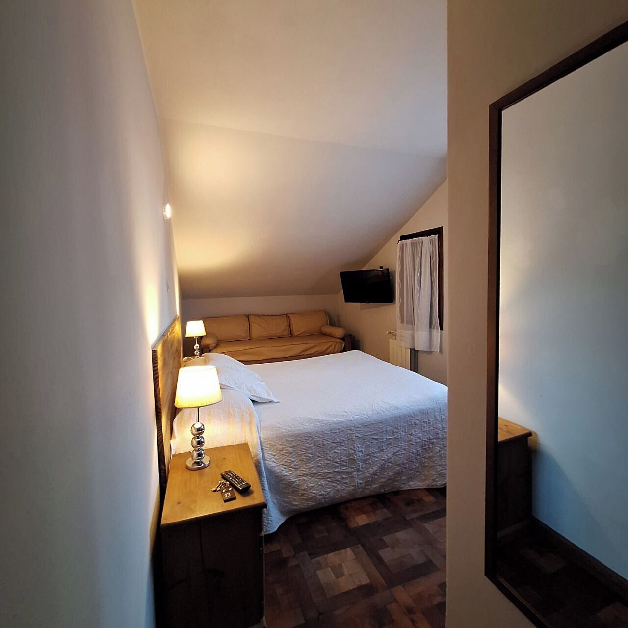 A small, cozy room with a bed, a bedside table with a lamp, a TV mounted on the wall, and a window with curtains.