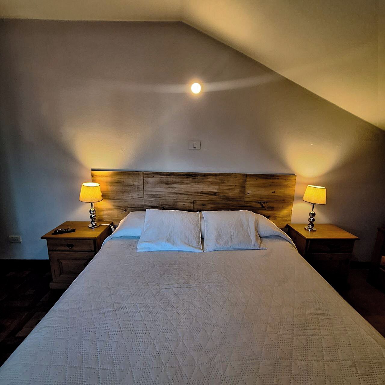 Un dormitorio con techo inclinado, cabecera de madera, dos lámparas de noche, cama con ropa de cama blanca y mesita de noche con un control remoto.