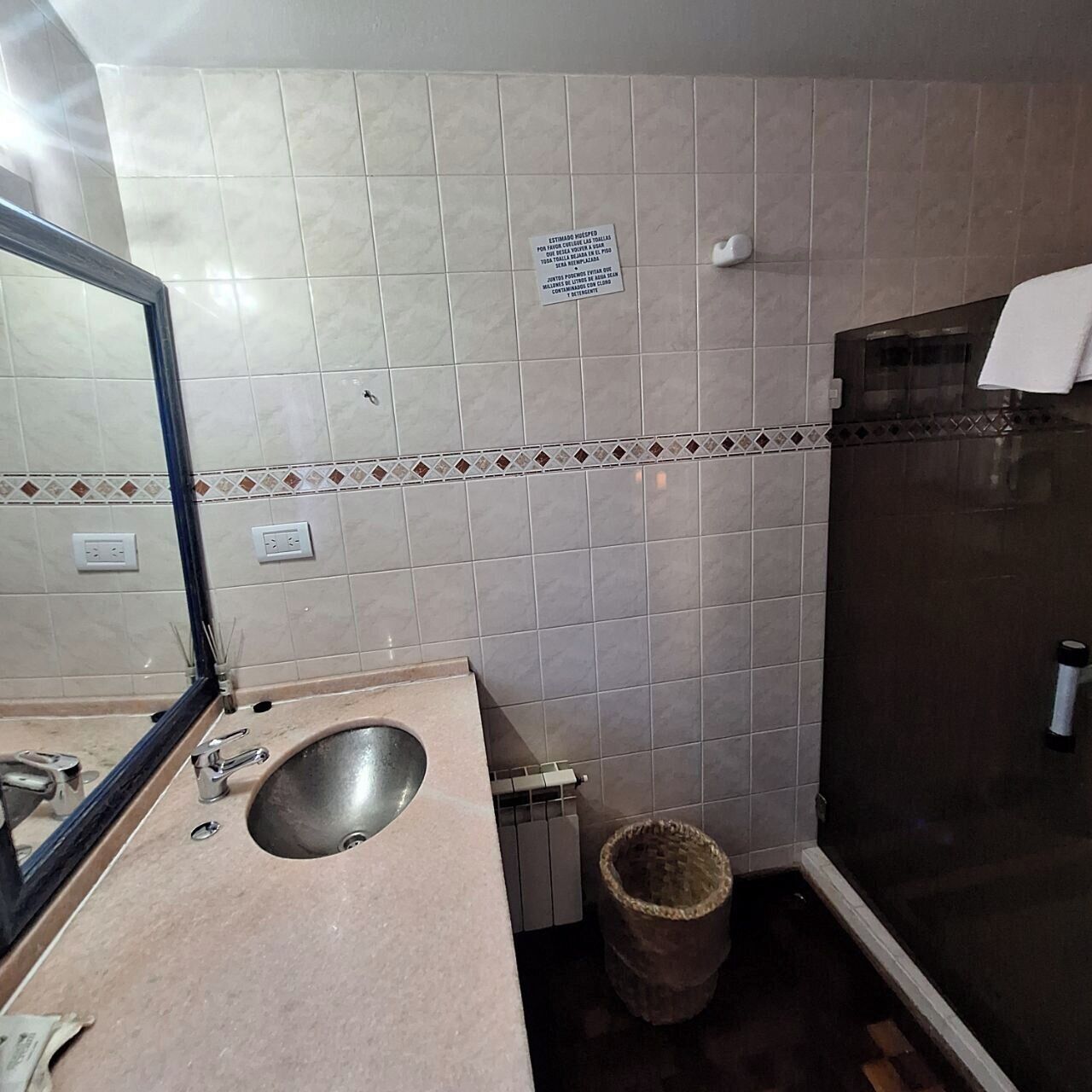 A bathroom with a sink, mirror, tiled walls, a trash bin, and a shower area.