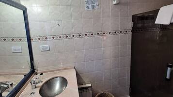 A bathroom with a sink, mirror, tiled walls, a trash bin, and a shower area.