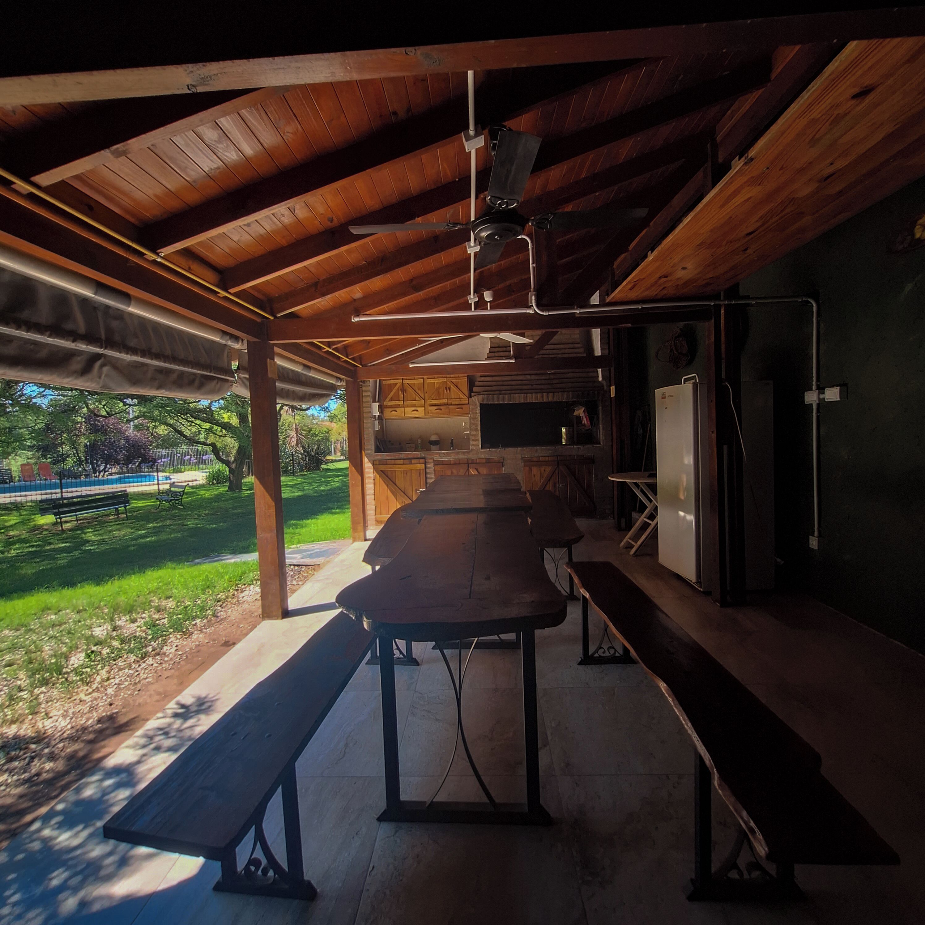 Un porche techado al aire libre con una mesa larga y bancos, un refrigerador y vistas a un jardín.