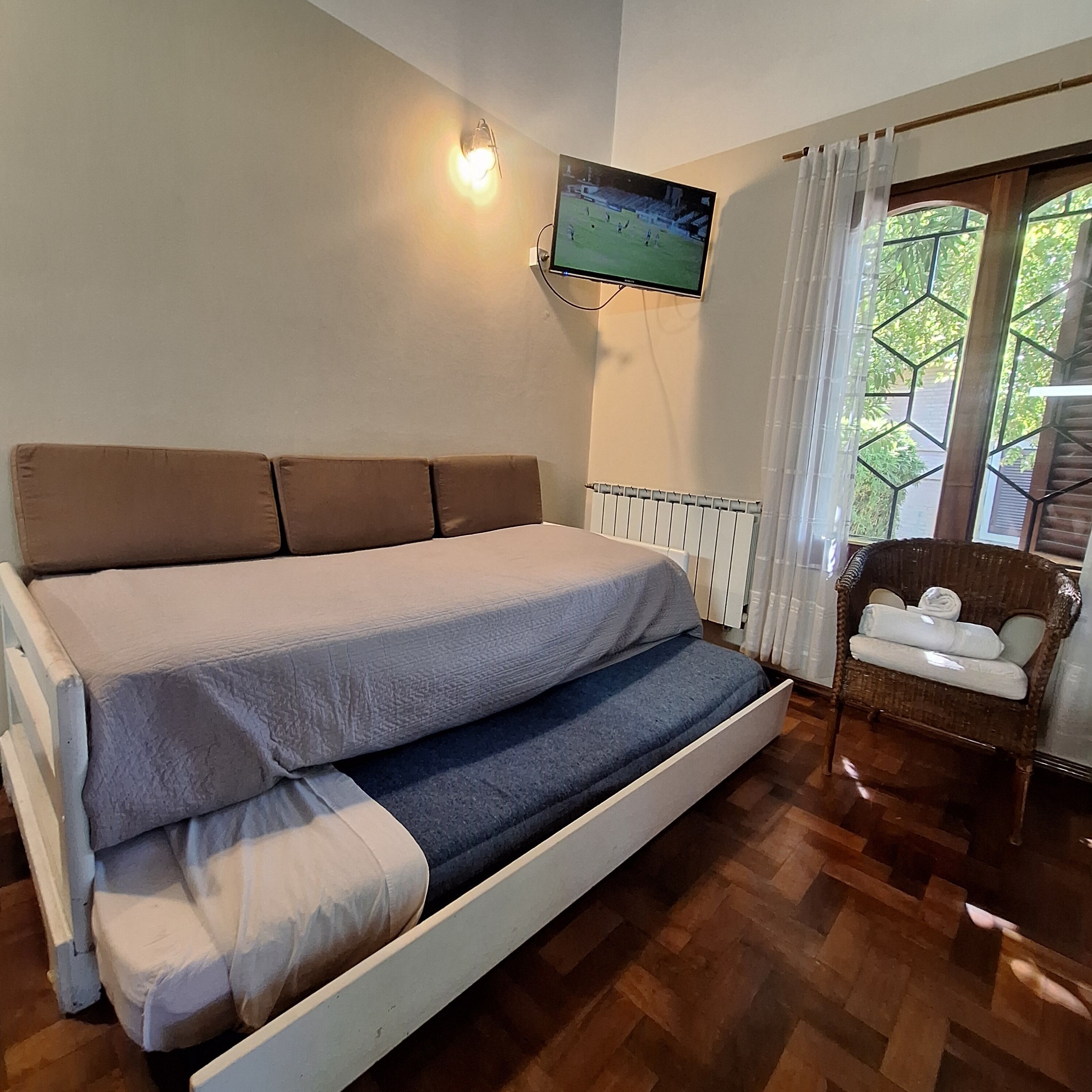 Ein Zimmer mit einem Bett, einem an der Wand befestigten Fernseher, einem Sessel und einem Fenster mit Vorhängen.