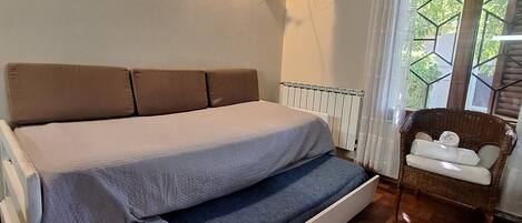 Ein Zimmer mit einem Bett, einem an der Wand befestigten Fernseher, einem Sessel und einem Fenster mit Vorhängen.
