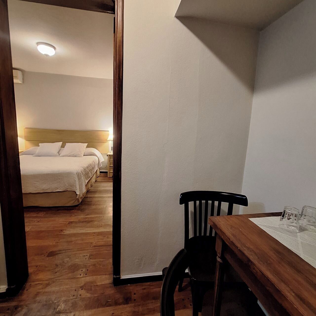 Una habitación de hotel con piso de madera, cama con ropa blanca, una mesa de madera con silla y una mesita de noche.