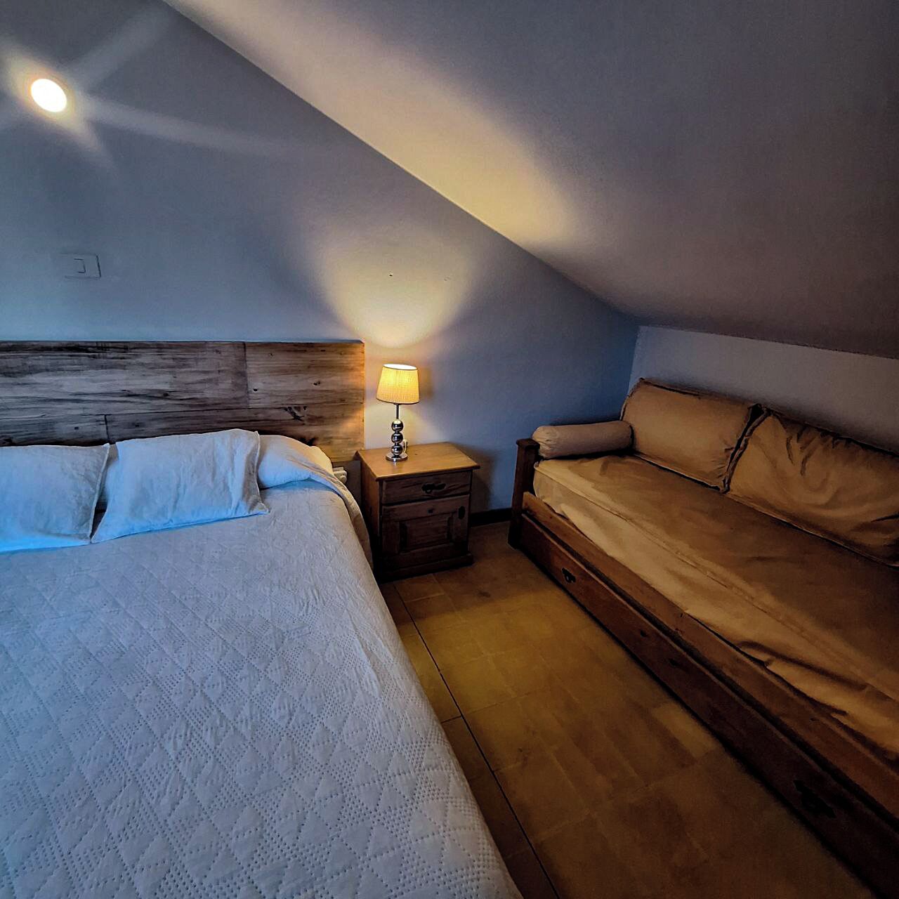 Un dormitorio con cabecera de madera, una mesita de noche con lámpara, una cama con colcha y un sofá.
