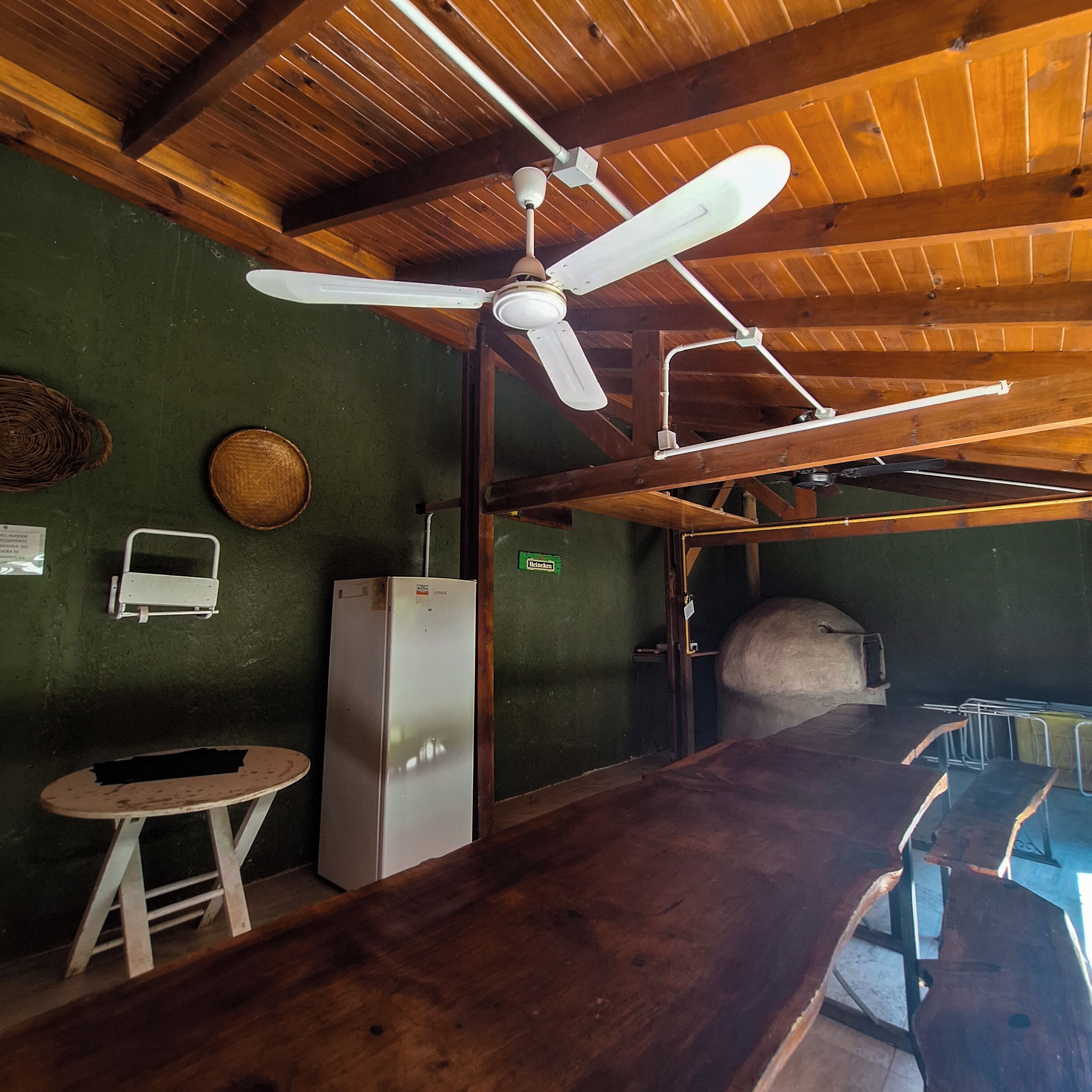 A rustic kitchen with a wooden ceiling, a ceiling fan, a refrigerator, a large oven, and a bar area with stools.