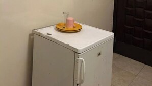 Un petit réfrigérateur blanc, surmonté d’une tasse rose, un tableau encadré représentant des fleurs accroché au mur, et une porte sombre ornée d’un motif géométrique.