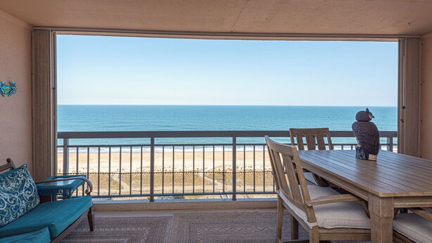 A balcony with a view of the beach and ocean, a wooden table, a wicker sofa, and a decorative owl statue. - Fountainhead-Oceanfront Retreat w/ Expansive Private Balcony (Ocean City)