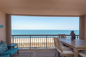 Un balcón con vista a la playa y al océano, una mesa de madera, un sofá de mimbre y una estatua decorativa de un búho.