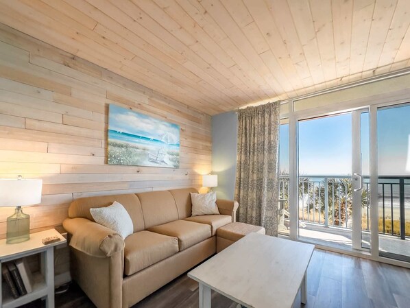 A room with a wooden ceiling, a beige sofa, a white table, a painting of a beach, and a balcony with a view. - Roxanne Towers 303 (Myrtle Beach)