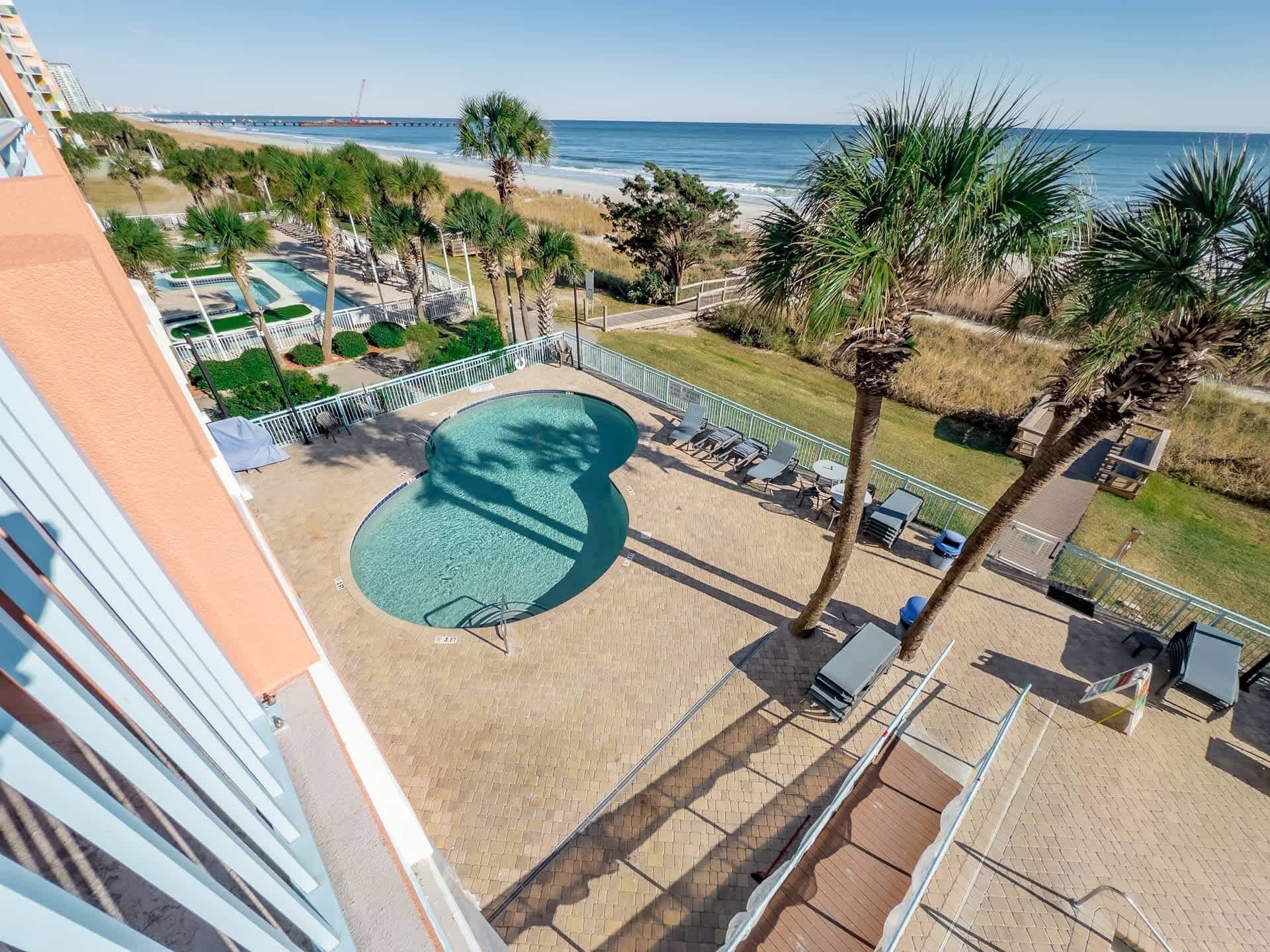 A pool area with a view of the beach and ocean, surrounded by palm trees and lounge chairs.