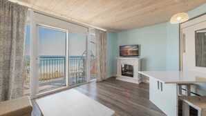 Een kamer met een houten plafond, een open haard, een televisie erboven en een balkon met uitzicht op het strand.