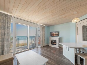 A room with a wooden ceiling, a fireplace, a TV mounted above it, and a balcony with a view of the beach. - Roxanne Towers 303 (Myrtle Beach)
