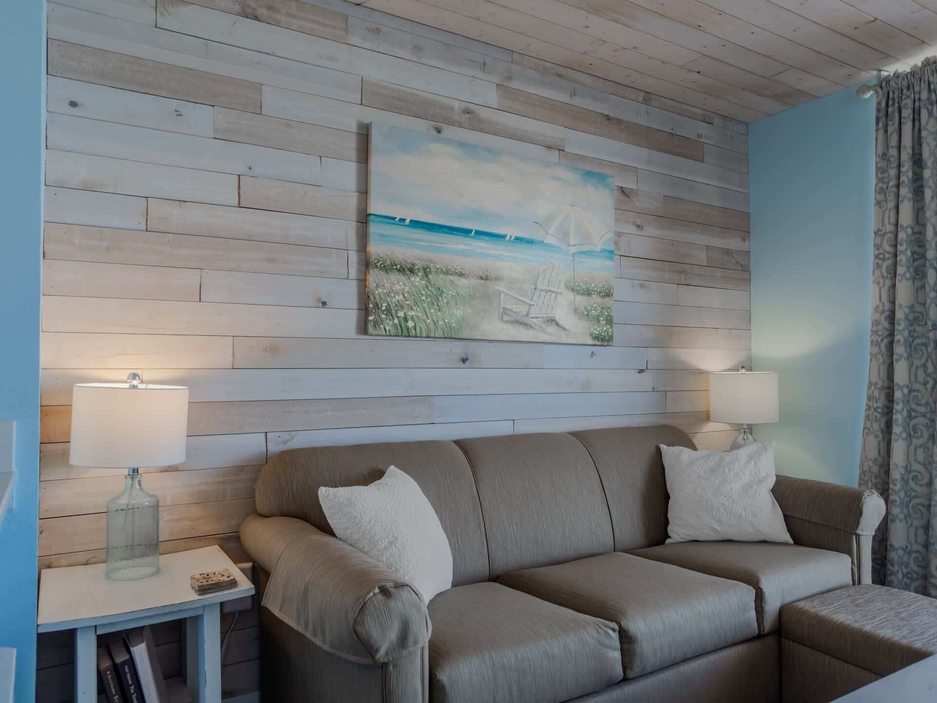 Una habitación con pared de madera, un cuadro con temática marina, un sofá con cojines y dos lámparas de noche.