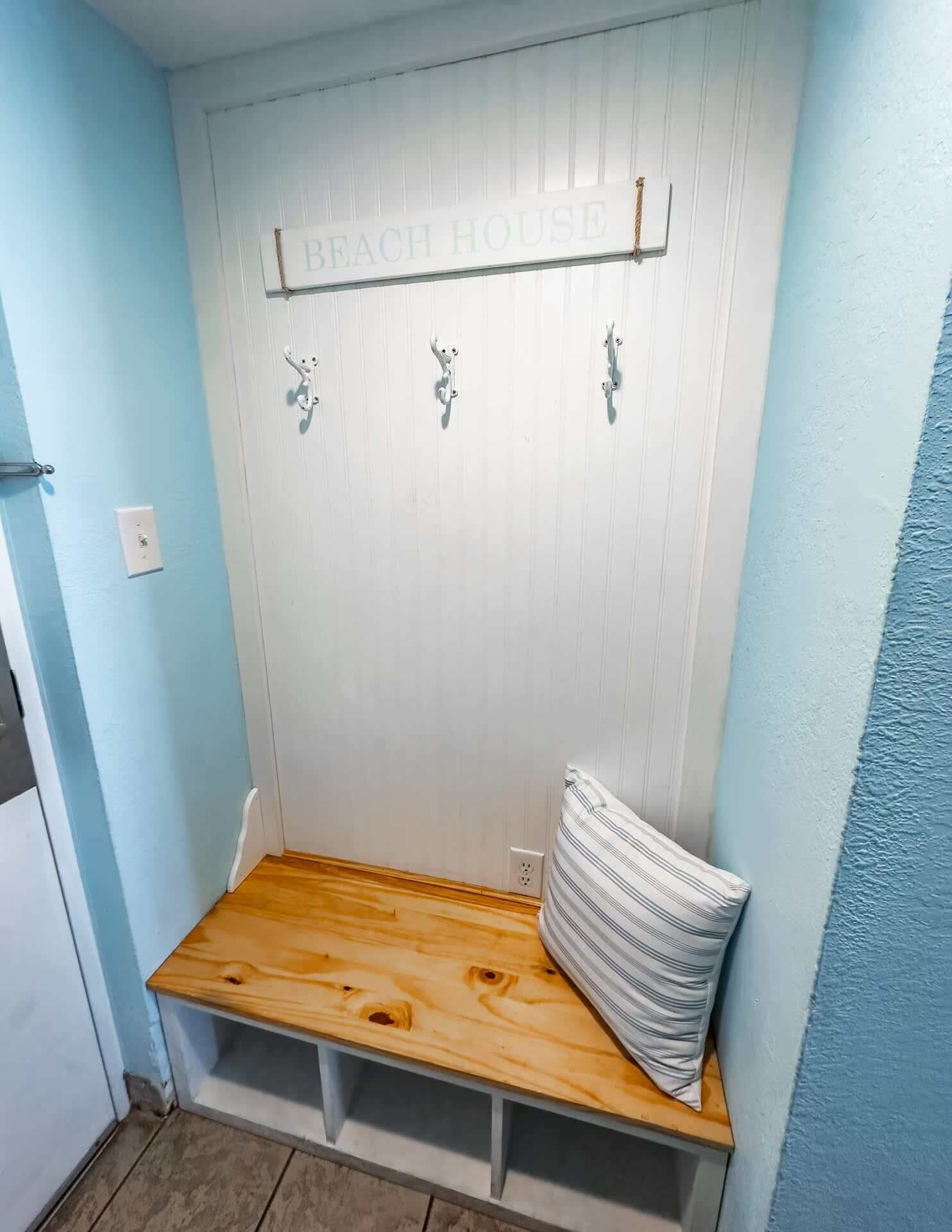 Ein Zimmer mit einer hölzernen Bank, einer weißen Wand mit dem Schild 'BEACH HOUSE' und einem kleinen weißen Kissen auf der Bank.
